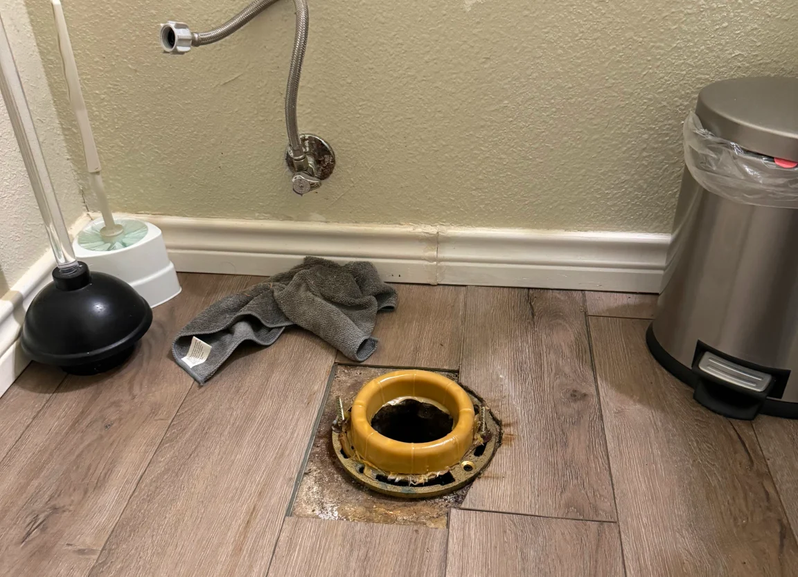 Exposed toilet flange with a yellow wax ring on a wood-look floor, with a disconnected supply line on the wall.
